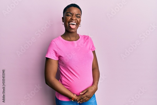 Young african american woman expecting a baby  touching pregnant belly sticking tongue out happy with funny expression. emotion concept.