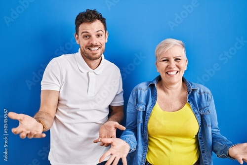 Young brazilian mother and son standing over blue background smiling cheerful with open arms as friendly welcome, positive and confident greetings