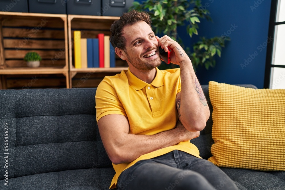 Young hispanic man talking on the smartphone sitting on sofa at home
