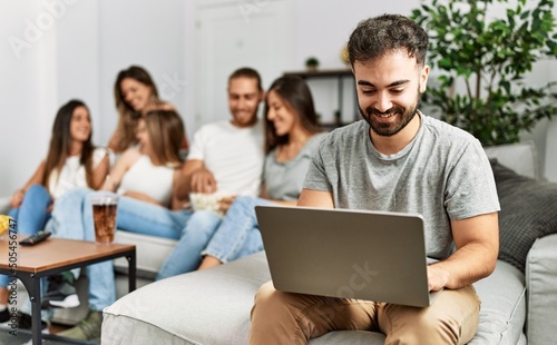Group of young friends speaking sitting on the sofa. Man smilimg happy and using laptop at home.