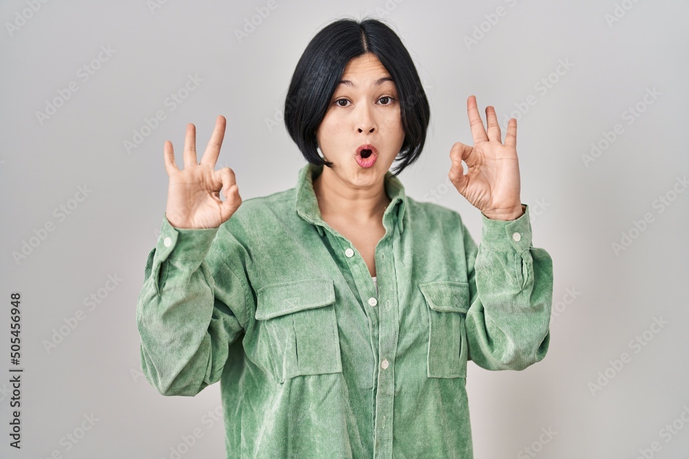 Young asian woman standing over white background looking surprised and shocked doing ok approval symbol with fingers. crazy expression Young asian woman standing over white background looking surprised and shocked doing ok approval symbol with fingers. crazy expression