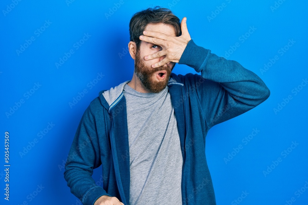 Caucasian man with beard wearing casual sweatshirt peeking in shock covering face and eyes with hand, looking through fingers with embarrassed expression. Caucasian man with beard wearing casual sweatshirt peeking in shock covering face and eyes with hand, looking through fingers with embarrassed expression.