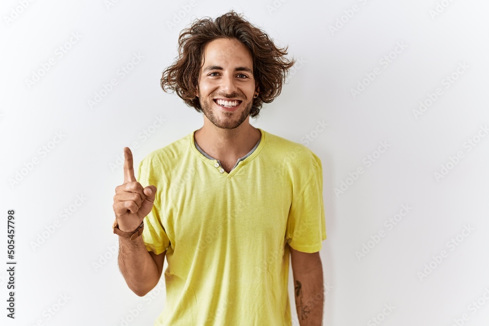Young hispanic man standing over isolated background showing and pointing up with finger number one while smiling confident and happy. Young hispanic man standing over isolated background showing and pointing up with finger number one while smiling confident and happy.