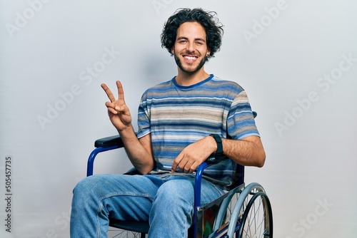 Handsome hispanic man sitting on wheelchair showing and pointing up with fingers number two while smiling confident and happy.