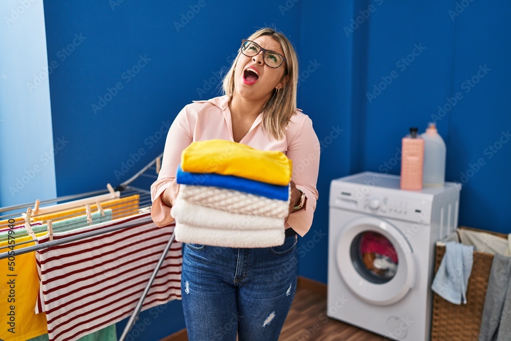Young hispanic woman holding folded laundry after ironing angry and mad screaming frustrated and furious, shouting with anger looking up.