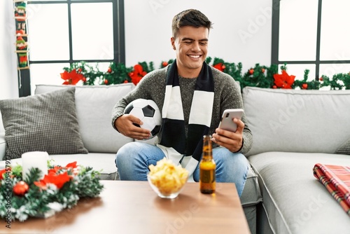 Young hispanic man supporting soccer game using smartphone sitting on sofa by christmas decor at home © Krakenimages.com