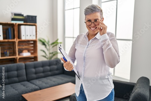 Middle age woman psychologist holding clipboard standing at pyschology center photo