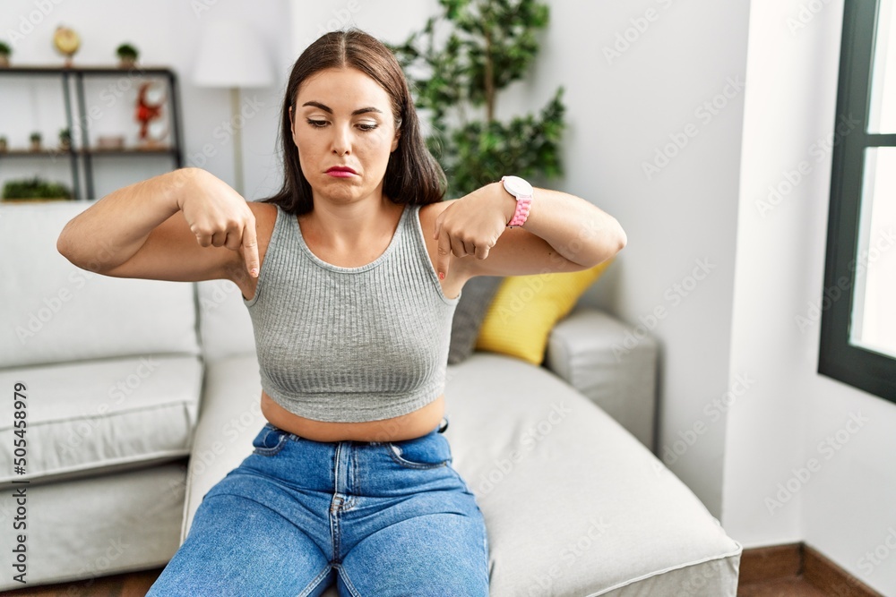 Young brunette woman sitting on the sofa at home pointing down looking sad and upset, indicating direction with fingers, unhappy and depressed.