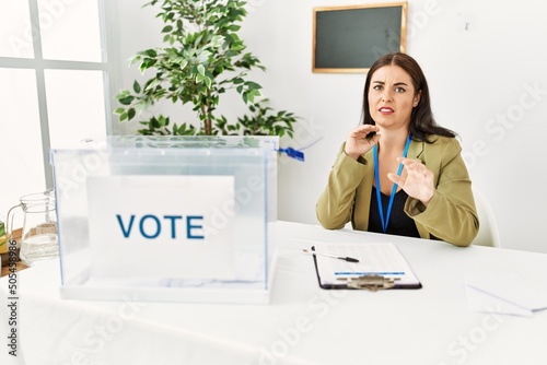 Young brunette woman sitting at election table with voting ballot disgusted expression, displeased and fearful doing disgust face because aversion reaction. with hands raised