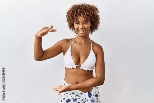 Young african american woman with curly hair wearing bikini gesturing with hands showing big and large size sign, measure symbol. smiling looking at the camera. measuring concept.