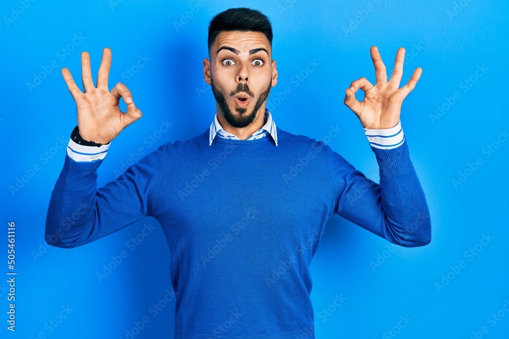Young hispanic man with beard wearing casual blue sweater looking surprised and shocked doing ok approval symbol with fingers. crazy expression Young hispanic man with beard wearing casual blue sweater looking surprised and shocked doing ok approval symbol with fingers. crazy expression