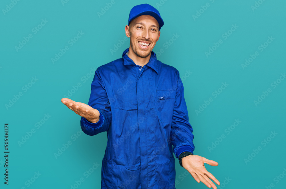 Bald man with beard wearing builder jumpsuit uniform smiling cheerful with open arms as friendly welcome, positive and confident greetings Bald man with beard wearing builder jumpsuit uniform smiling cheerful with open arms as friendly welcome, positive and confident greetings