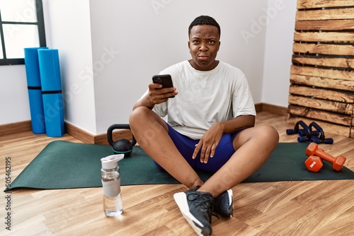 Young african man sitting on training mat at the gym using smartphone making fish face with lips  crazy and comical gesture. funny expression.