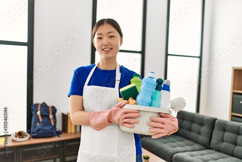 Young chinese housewife holding cleaning products standing at home.