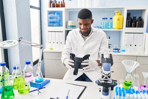Young african american man wearing scientist uniform holding virtual reality glasses at laboratory