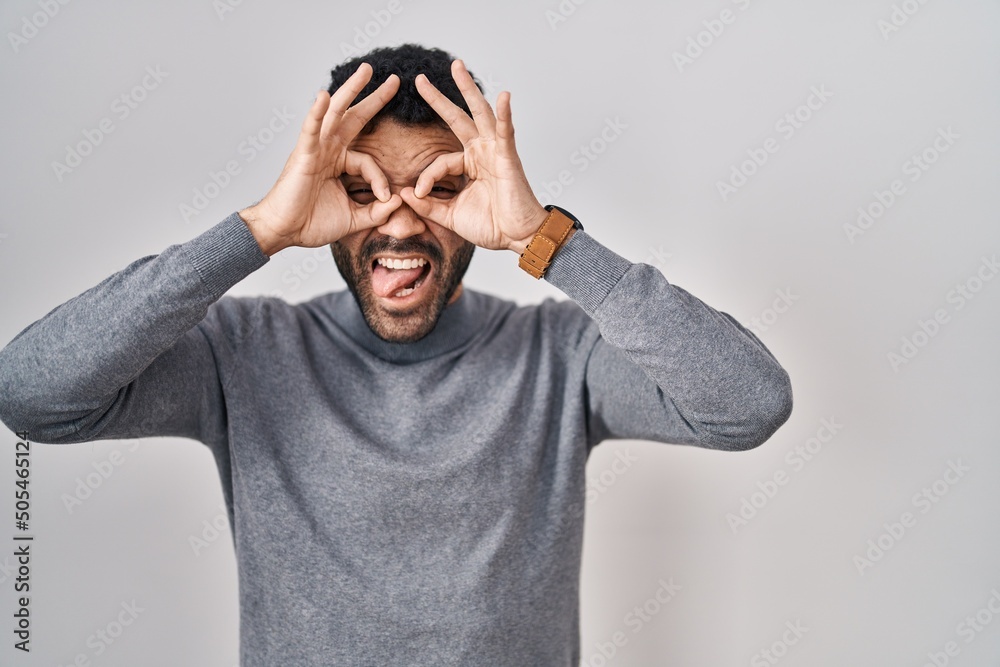 Hispanic man with beard standing over white background doing ok gesture like binoculars sticking tongue out, eyes looking through fingers. crazy expression. Hispanic man with beard standing over white background doing ok gesture like binoculars sticking tongue out, eyes looking through fingers. crazy expression.