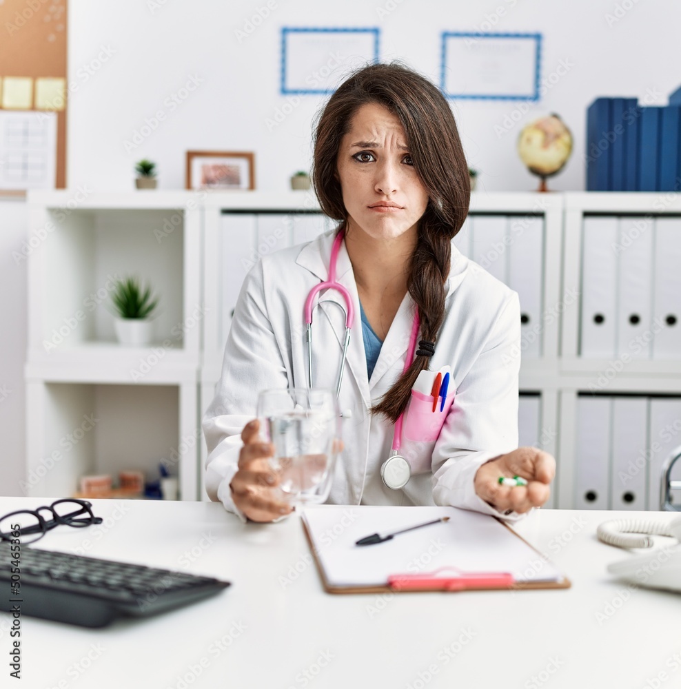 Young doctor woman holding glass of water and prescription pills skeptic and nervous, frowning upset because of problem. negative person. Young doctor woman holding glass of water and prescription pills skeptic and nervous, frowning upset because of problem. negative person.