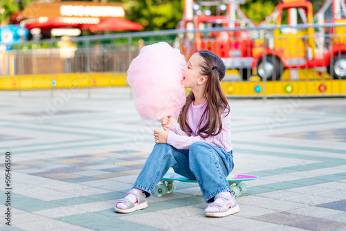 a child girl in an amusement park in the summer eats cotton candy on a skateboard and smiles with happiness near the carousels, the concept of summer holidays and school holidays photo