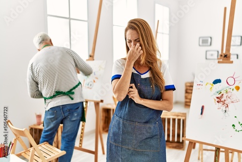 Hispanic woman wearing apron at art studio smelling something stinky and disgusting, intolerable smell, holding breath with fingers on nose. bad smell
