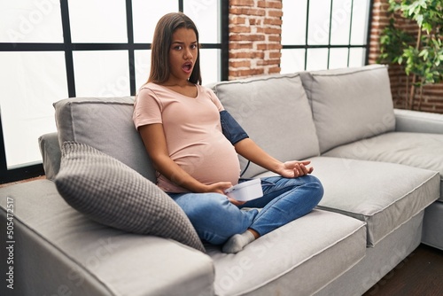 Young pregnant woman using blood pressure monitor sitting on the sofa in shock face, looking skeptical and sarcastic, surprised with open mouth