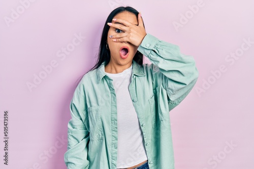 Beautiful hispanic woman with nose piercing wearing casual green jacket peeking in shock covering face and eyes with hand, looking through fingers afraid © Krakenimages.com