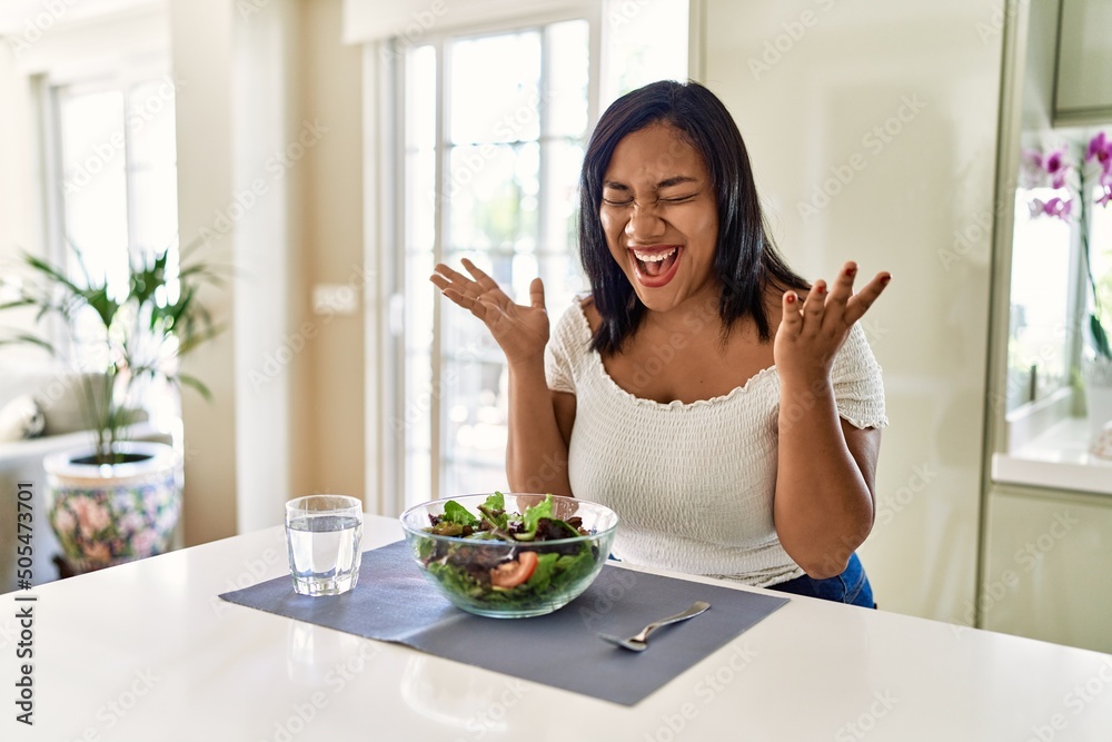 Young hispanic woman eating healthy salad at home celebrating mad and crazy for success with arms raised and closed eyes screaming excited. winner concept