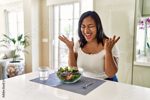Young hispanic woman eating healthy salad at home celebrating mad and crazy for success with arms raised and closed eyes screaming excited. winner concept