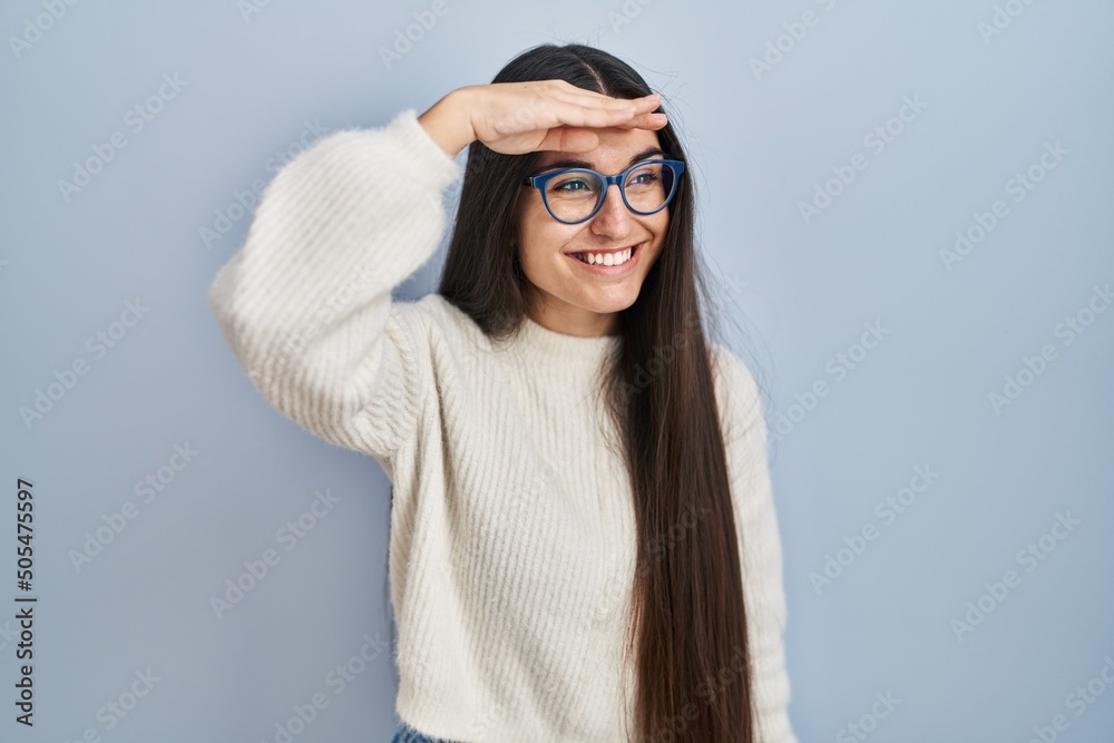 Young hispanic woman wearing casual sweater over blue background very happy and smiling looking far away with hand over head. searching concept.