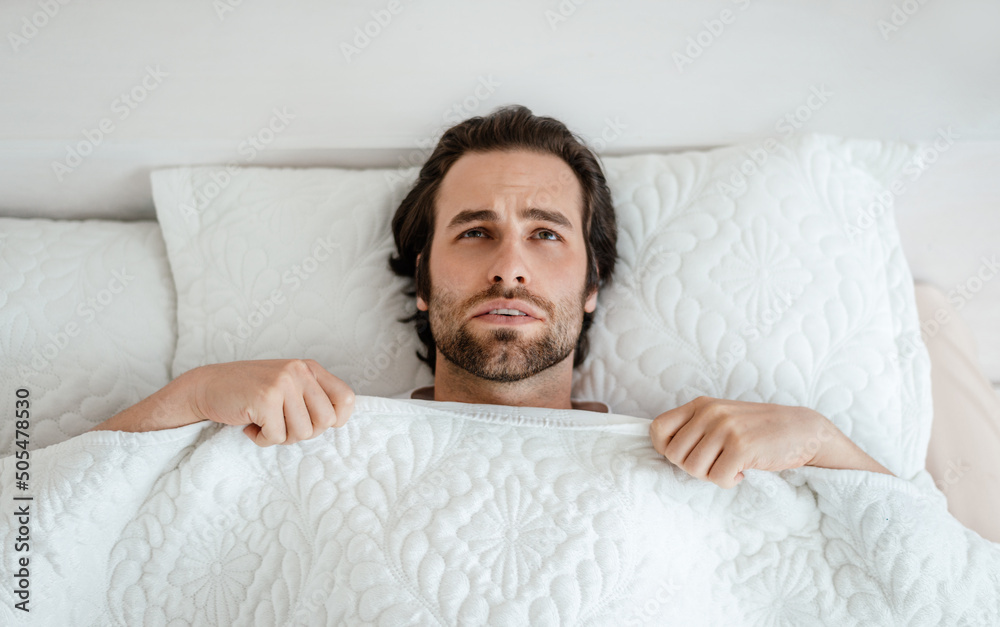 Unhappy millennial caucasian man with stubble wake up, feeling bad, lying on white bed in bedroom interior Unhappy millennial caucasian man with stubble wake up, feeling bad, lying on white bed in bedroom interior