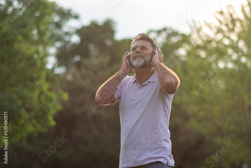 Happy mature bearded man listening to music. Senior stylish bearded man listening to music in the city park. Relaxed man breathing fresh air and meditating listening audio guide with headphones.