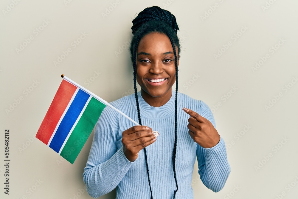 African american woman with braided hair holding gambia flag smiling happy pointing with hand and finger - obrazy, fototapety, plakaty 