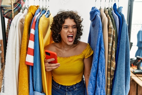 Young hispanic woman searching clothes on clothing rack using smartphone angry and mad screaming frustrated and furious, shouting with anger. rage and aggressive concept.