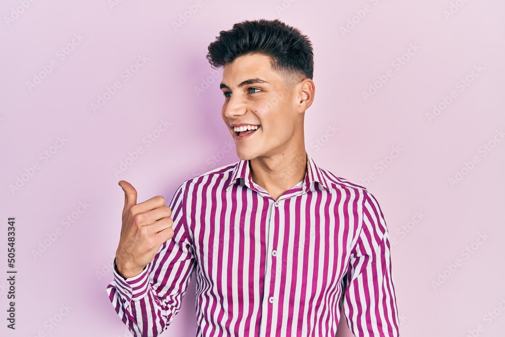 Young hispanic man wearing casual clothes smiling with happy face looking and pointing to the side with thumb up. Young hispanic man wearing casual clothes smiling with happy face looking and pointing to the side with thumb up.