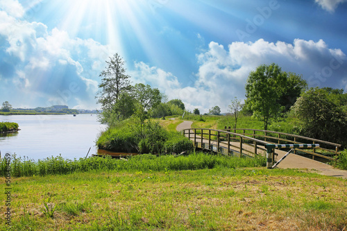 Beautiful dutch rural landscape, cycle track along river maas over old wood bridge, green trees, sun rays, blue cloudy sky - Limburg, South Holland photo