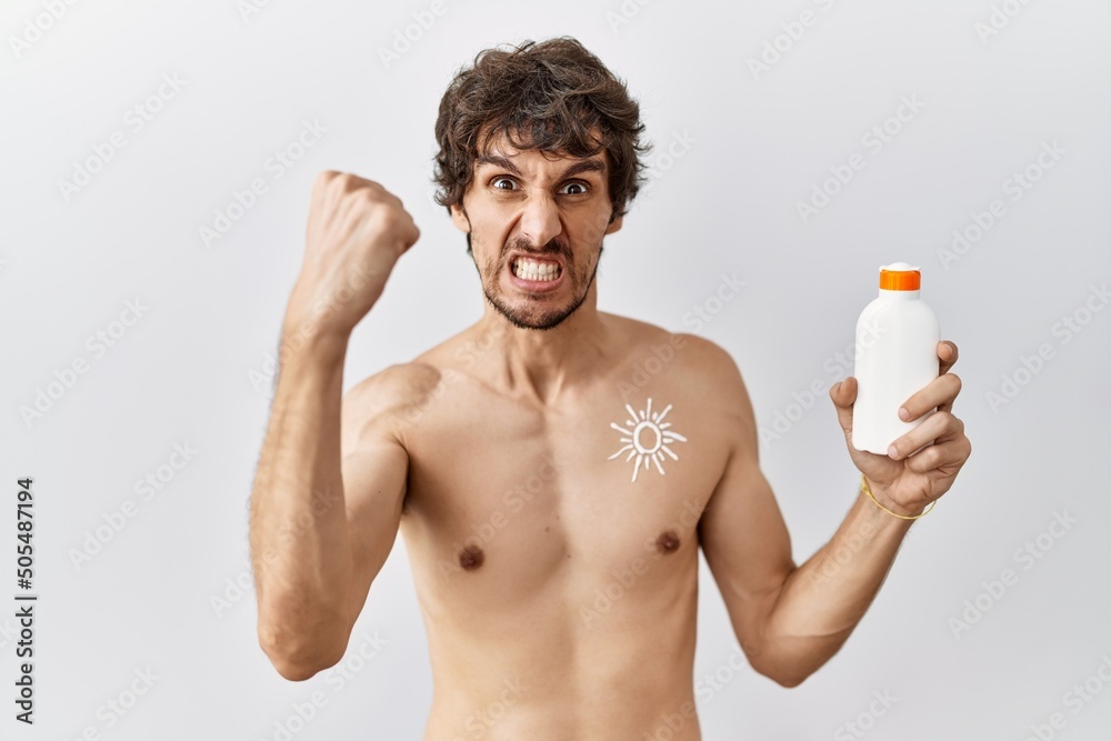 Fototapeta premium Young hispanic man standing shirtless holding sunscreen lotion angry and mad raising fist frustrated and furious while shouting with anger. rage and aggressive concept.