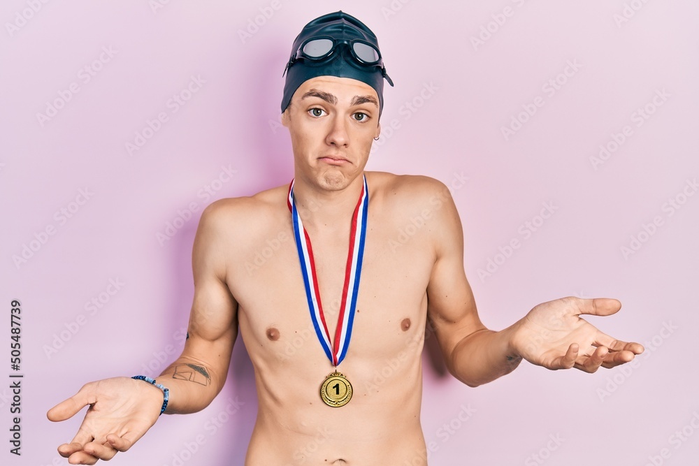Young hispanic man wearing swimmer glasses and gold medal clueless and confused expression with arms and hands raised. doubt concept.