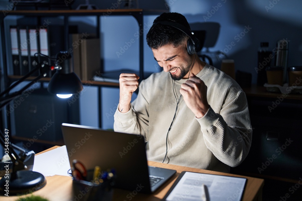 Young handsome man working using computer laptop at night very happy and excited doing winner gesture with arms raised, smiling and screaming for success. celebration concept.