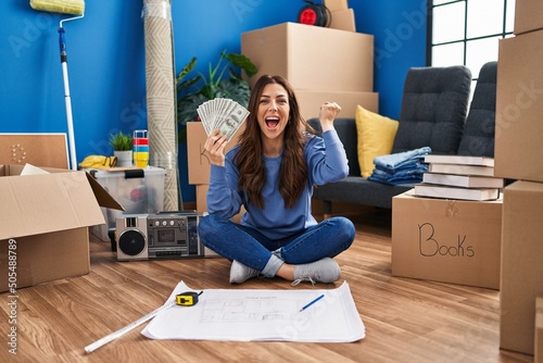 Young brunette woman sitting on the floor at new home holding money celebrating victory with happy smile and winner expression with raised hands photo