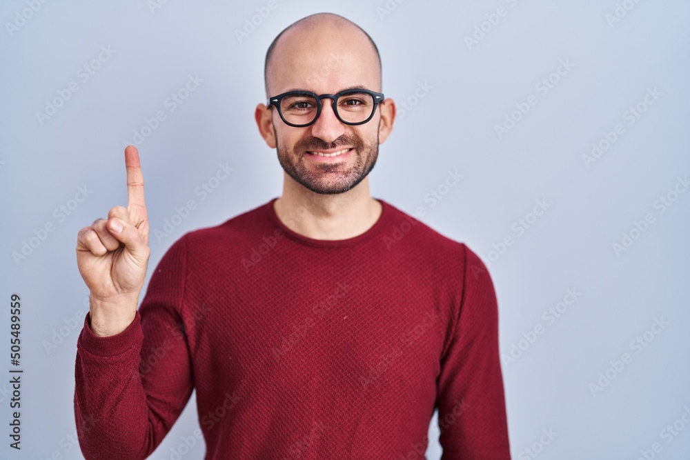 Φωτογραφία Young bald man with beard standing over white background wearing glasses showing and pointing up with finger number one while smiling confident and happy