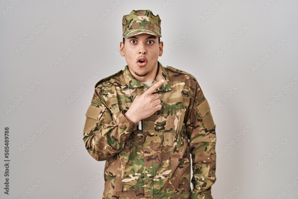 Young arab man wearing camouflage army uniform surprised pointing with finger to the side, open mouth amazed expression. Young arab man wearing camouflage army uniform surprised pointing with finger to the side, open mouth amazed expression.