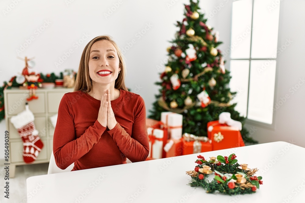 Caucasian young blonde woman sitting on the table by christmas tree praying with hands together asking for forgiveness smiling confident. Caucasian young blonde woman sitting on the table by christmas tree praying with hands together asking for forgiveness smiling confident.