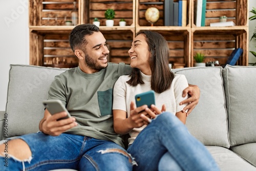 Young latin couple using smartphone sitting on the sofa at home.