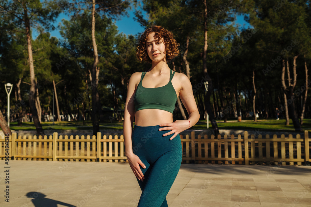 Beautiful young redhead woman wearing green sports bra and blue yoga pants poses to camera. Sportive woman looking at the camera while standing on city park, outdoors.