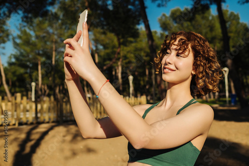 Photo of sporty trainer coach lady recording video selfie smart device on city park, outdoors. Young redhead woman taking selfie with her phone. Outdoor sports and healthy life concepts.