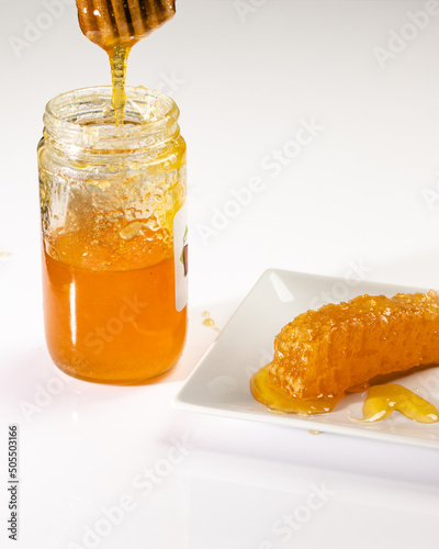 Closeup of honey dripping from honey dipper with a honeycomb in a dish on the side all sitting on a white plexiglas background