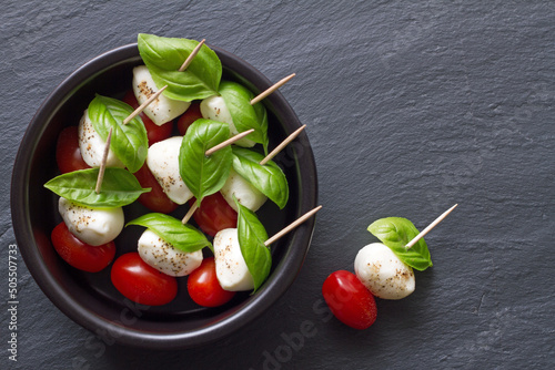Caprese sticks with mozzarella, tomatoes and basil leaves in bowl on black background