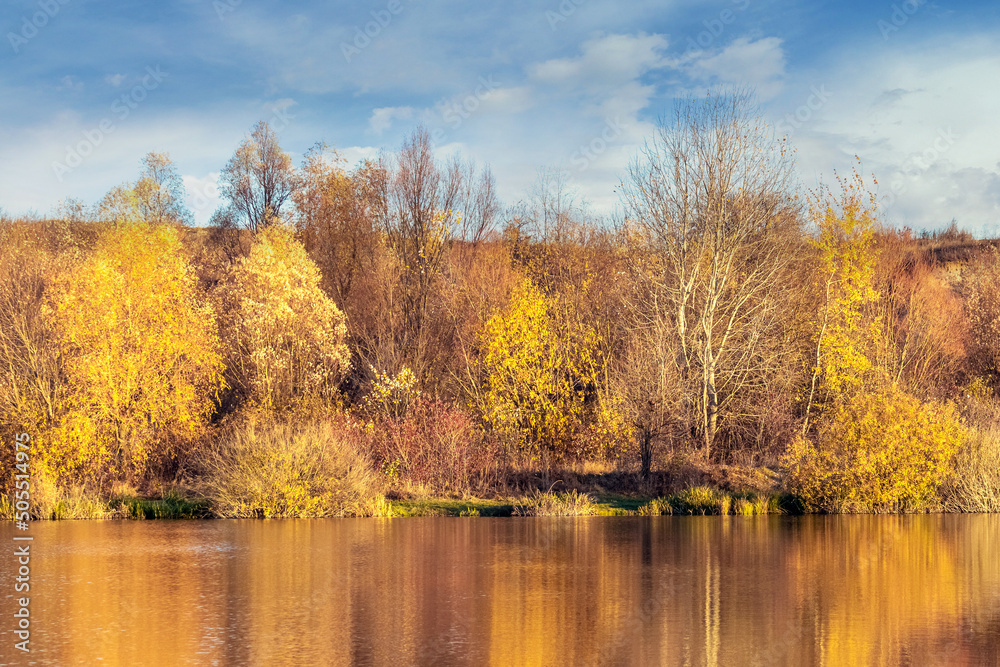 Picturesque autumn landscape with yellow trees over the river and reflection of trees in the water on a sunny day. Golden autumn
