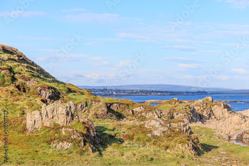 A scenic seaside view with rock and grass in the foreground and coastline village and blue sea in the background under a majestic blue sky and some white clouds