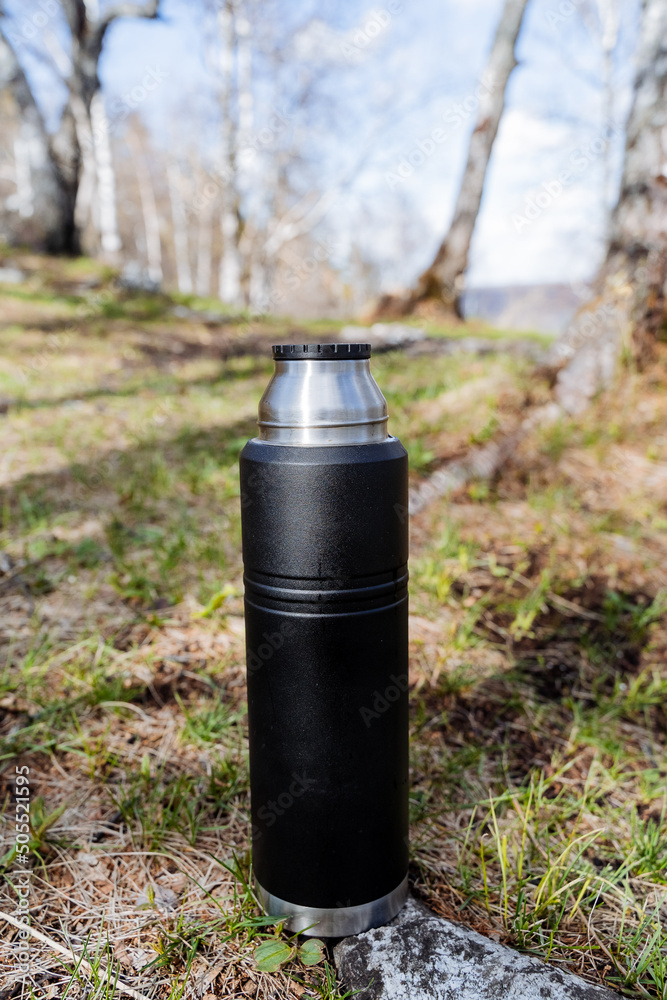 Thermos for tea stands on a stone against the background of the forest, black steel bottle, durable metal vacuum bottle, hot drinks, camping thermo cup, brewed coffee Thermos for tea stands on a stone against the background of the forest, black steel bottle, durable metal vacuum bottle, hot drinks, camping thermo cup, brewed coffee