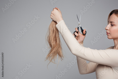 Studio portrait of blonde female cutting her own hair, hairstyle and haircut, lock of hair, scissors in hand, plain background, isolated, changes and hair treatment concept, lifestyle beauty and style